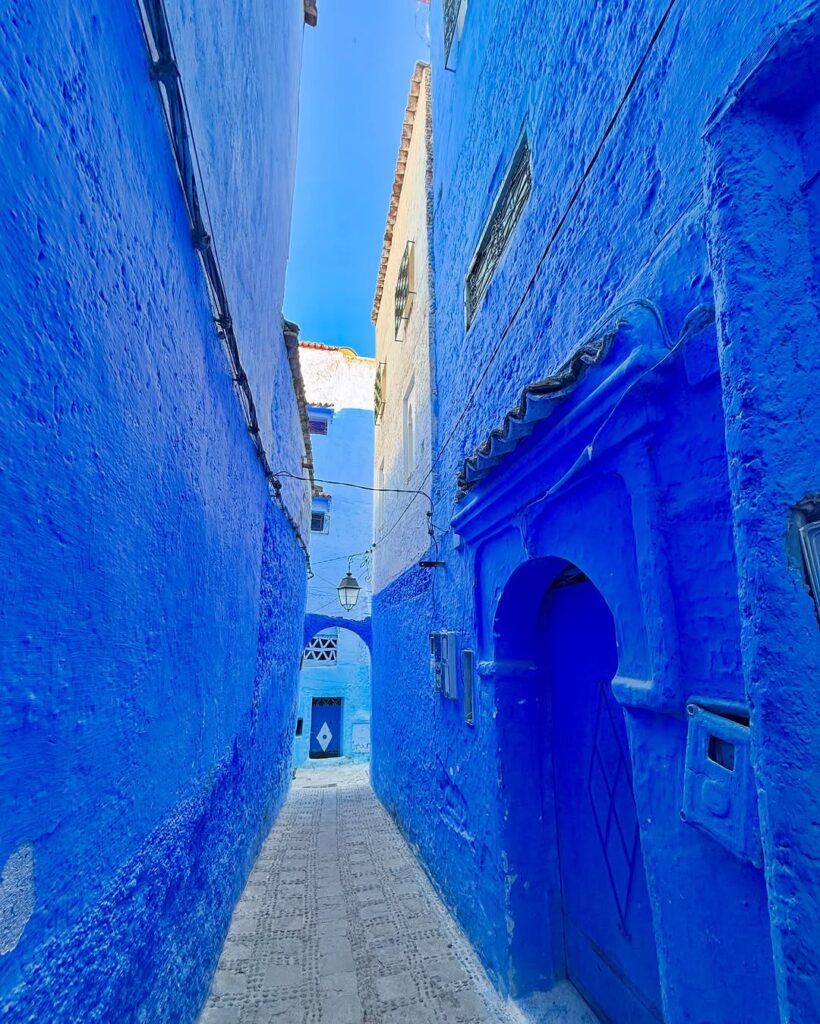 Narrow, vibrant blue-washed alleyway featuring a cobbled street and a traditional arched doorway in the picturesque historic medina of Chefchaouen, Morocco.