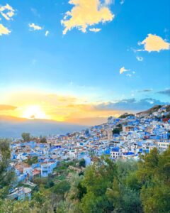Panoramic sunset view over the iconic blue-washed hillside buildings of Chefchaouen, Morocco, with golden sunlight breaking through the mountains.