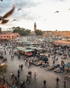 Busy market square in Marrakesh featuring crowds, stalls, terracotta architecture, and the Koutoubia Mosque minaret.
