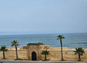 Coastal exterior of Restaurant Borj Eddar in Morocco, showcasing a traditional Moroccan arched entrance and stone walls bordered by palm trees overlooking the ocean waves.