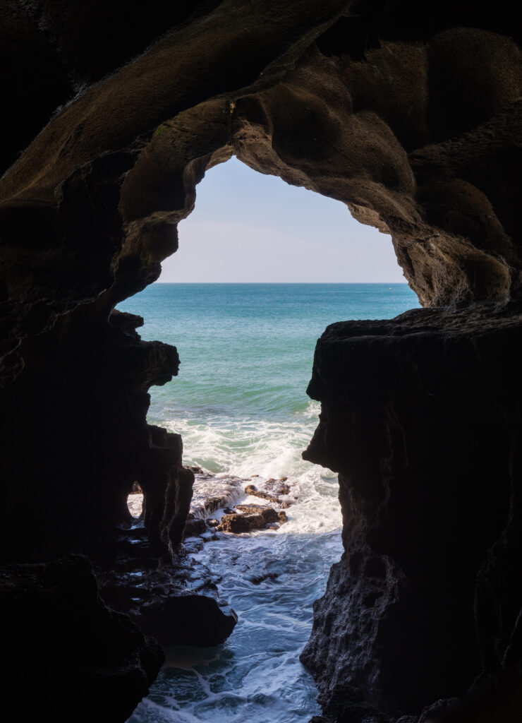 Scenic view looking out from inside the historic Cave of Hercules in Tangier, Morocco, through its unique ocean-facing opening shaped remarkably like the continent of Africa, with waves crashing against rocky shores and a clear blue sea.