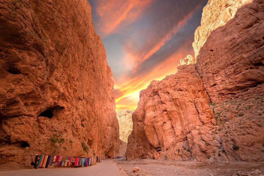 Breathtaking sunrise view over the towering red limestone cliffs of Todgha Gorge in Morocco, illuminating the narrow canyon path lined with colorful traditional Berber carpets under vibrant orange and pink clouds.