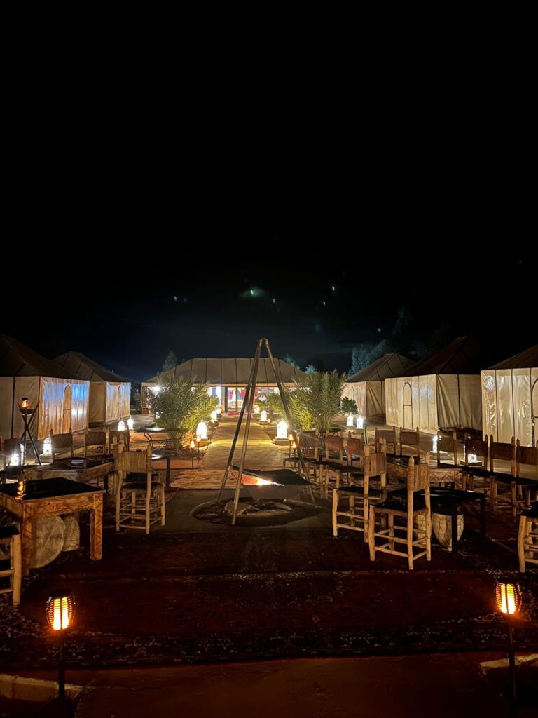 Luxury nighttime desert glamping camp featuring glowing white canvas tents, warm lantern lighting, traditional patterned rugs, and a central fire pit gathering area.