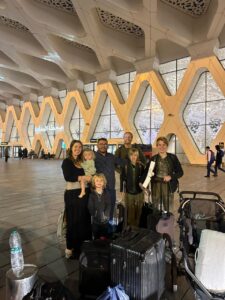 Smiling family posing with luggage inside the modern terminal of Marrakech Menara Airport in Morocco, highlighted by the striking contemporary architecture of repeating white geometric arches and glass panels.