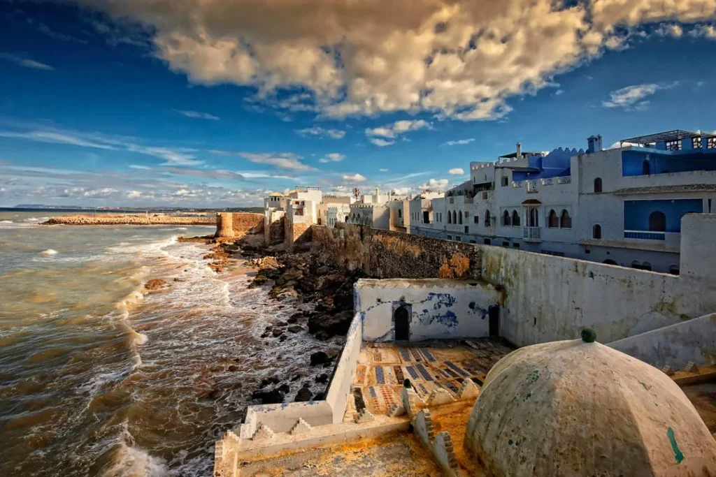 Scenic coastal view of the historic fortified ramparts and white-washed medina walls of Assilah, Morocco, with waves crashing against the rocky Atlantic shore under a dramatic cloudy blue sky.