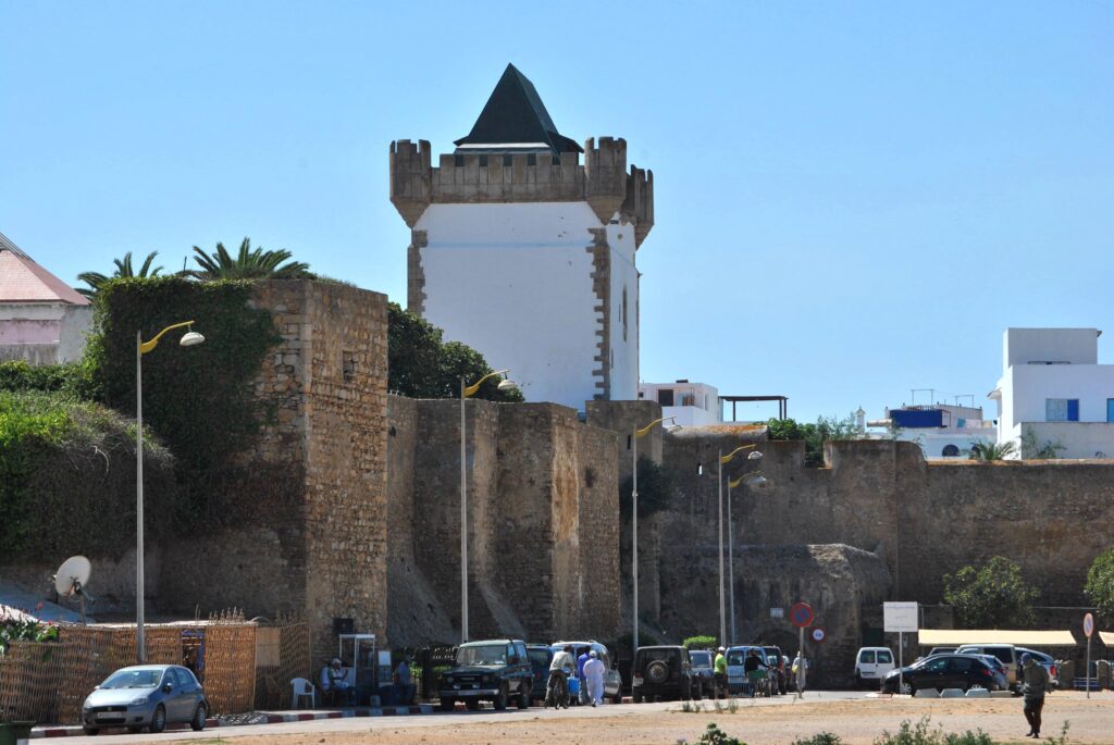Historic fortified stone city walls and defense towers of Assilah, Morocco, featuring a prominent white watchtower with a dark pointed roof under a clear blue sky.