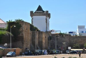 Historic fortified stone city walls and defense towers of Assilah, Morocco, featuring a prominent white watchtower with a dark pointed roof under a clear blue sky.