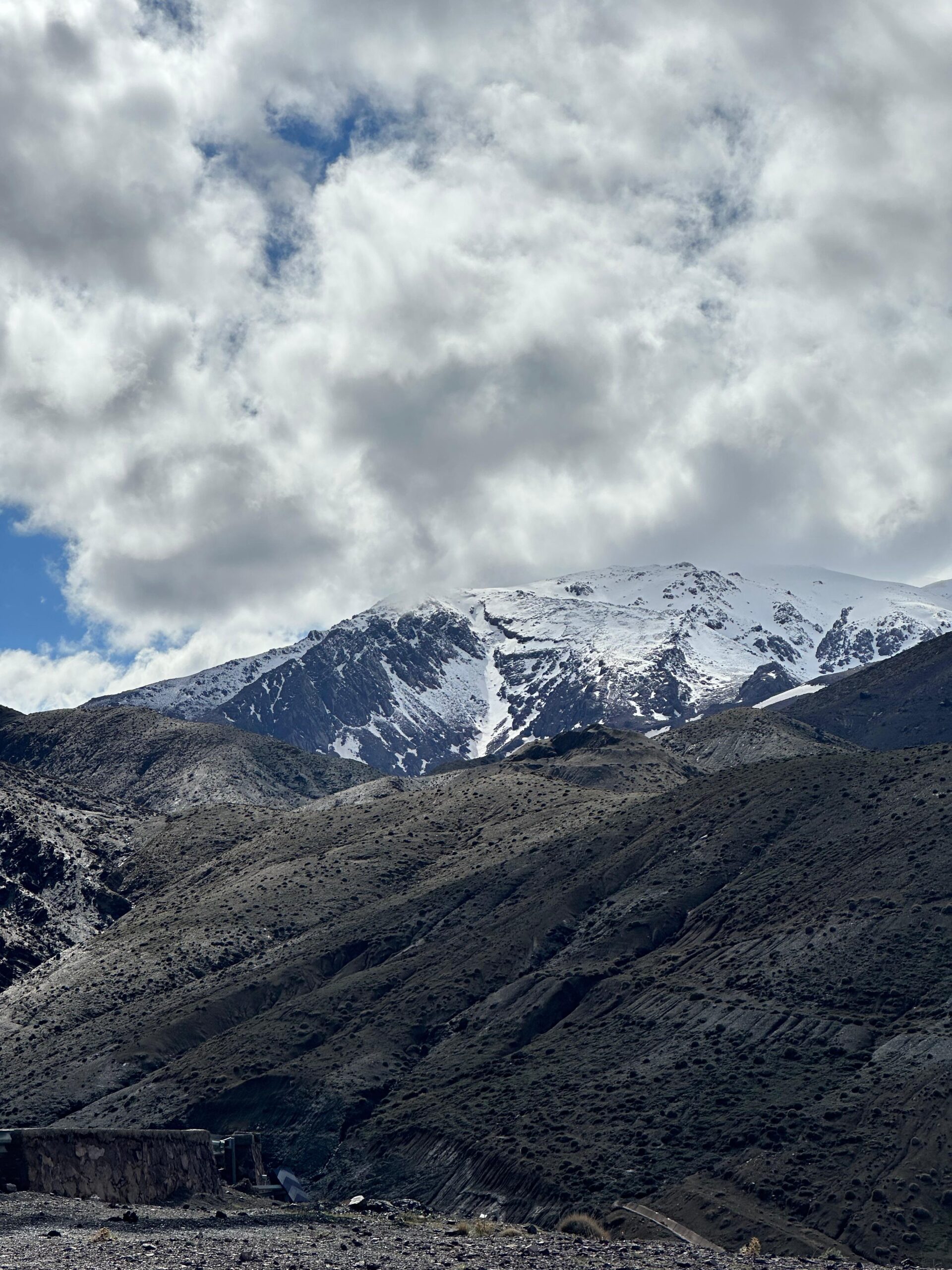 Rugged, barren slopes of the High Atlas Mountains in Morocco leading up to a dramatic snow-covered alpine peak under a dynamic, heavily clouded sky.