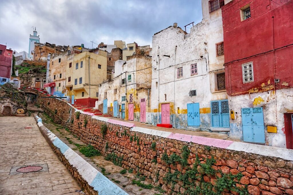 Street view of colorful, historic houses built into the hillside in the troglodyte village of Bhalil, Morocco, featuring stone walls and painted doors.
