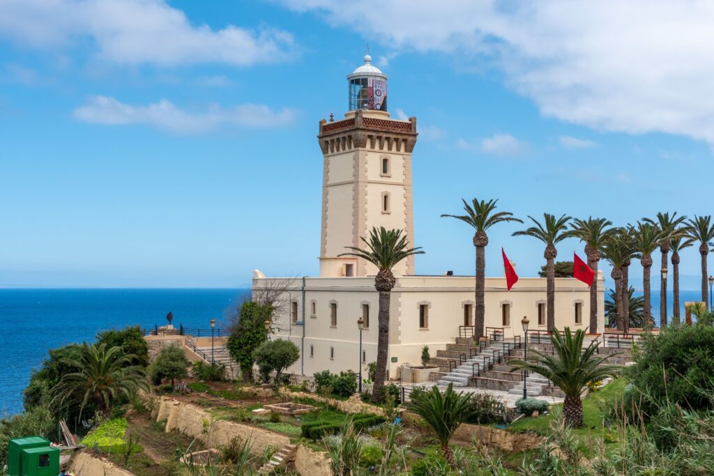 Scenic view of the historic Cap Spartel lighthouse standing proudly on a coastal cliff in Morocco, surrounded by palm trees, landscaped gardens, and flying Moroccan flags against a blue sky with ocean views.