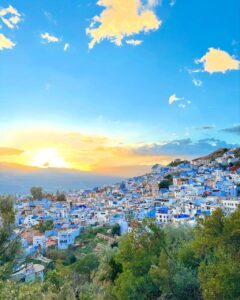 Panoramic sunset view of the famous blue city of Chefchaouen in Morocco, featuring blue and white hillside architecture nestled in the Rif Mountains under a vibrant glowing sky with golden clouds.