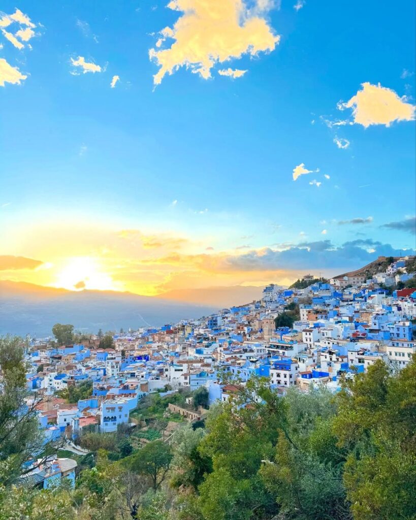 Panoramic sunset view of the famous blue city of Chefchaouen in Morocco, featuring blue and white hillside architecture nestled in the Rif Mountains under a vibrant glowing sky with golden clouds.