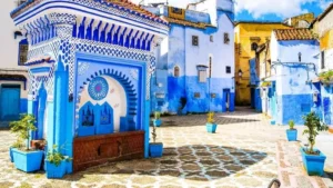 Ornate blue and white traditional public fountain in a sunny square of the blue city of Chefchaouen, Morocco, surrounded by blue-washed buildings, potted plants, and patterned stone paving.