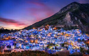 Stunning twilight panoramic view of the blue city of Chefchaouen, Morocco, illuminated by warm city lights against the dark silhouette of the Rif Mountains under a vibrant purple and orange sunset sky.