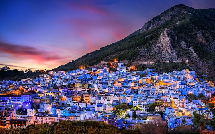 Stunning twilight panoramic view of the blue city of Chefchaouen, Morocco, illuminated by warm city lights against the dark silhouette of the Rif Mountains under a vibrant purple and orange sunset sky.