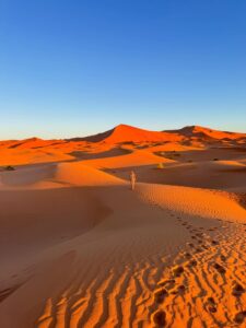 Person walking through large orange sand dunes in the desert at sunset, featuring rippled sand, deep shadows, and a clear blue sky.
