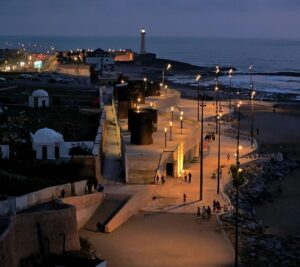 Illuminated seaside promenade at night along the Moroccan coast, featuring a historic lighthouse, modern curved walkways, and ancient stone fortress walls beside the Atlantic Ocean.