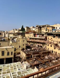 Panoramic view of the historic Chouara Tannery in the ancient medina of Fes, Morocco, featuring traditional stone dyeing vats filled with liquids and hides, surrounded by multi-story yellow buildings under a clear blue sky.