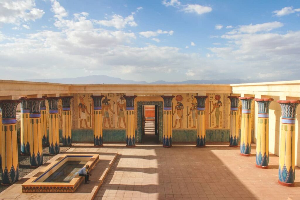 Egyptian-style film set at Atlas Film Studios in Ouarzazate, Morocco, featuring colorful columns, detailed wall murals depicting ancient Egyptian gods, and a courtyard pool under a bright sky.