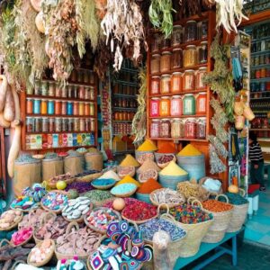 Colorful display at a traditional Moroccan spice shop in a medina souk, featuring perfectly shaped pyramids of vibrant spices, woven baskets of dried flowers, and hanging aromatic herbs.