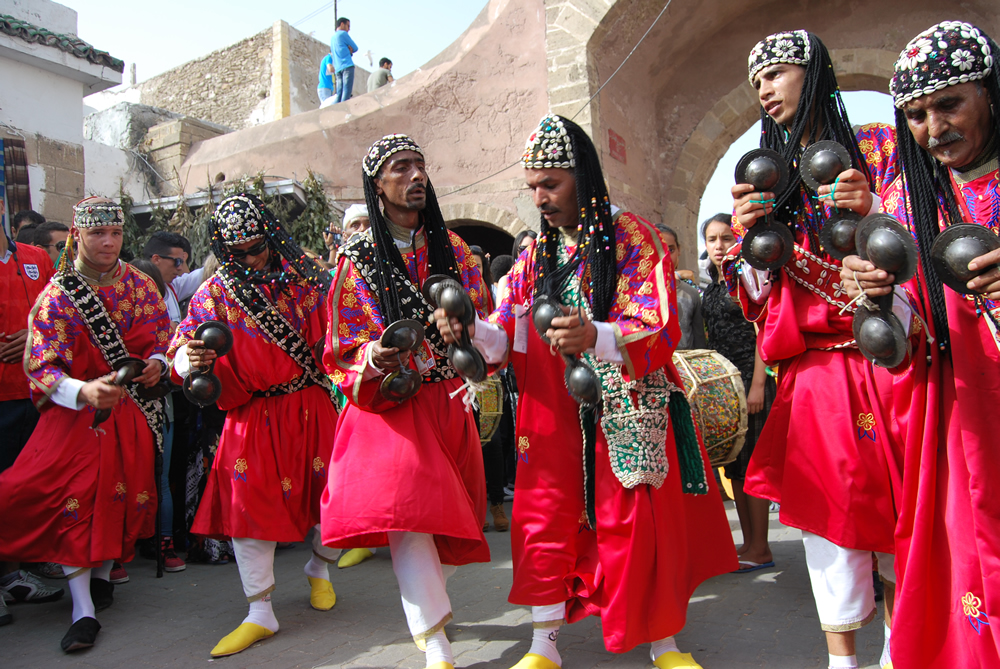 Traditional Moroccan Gnawa musicians wearing vibrant red embroidered robes and cowrie shell caps, playing metallic krakeb castanets during a lively cultural street performance.