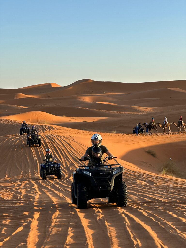 Desert adventure tourism featuring an ATV quad bike tour driving through orange sand dunes with a traditional camel caravan riding in the background.