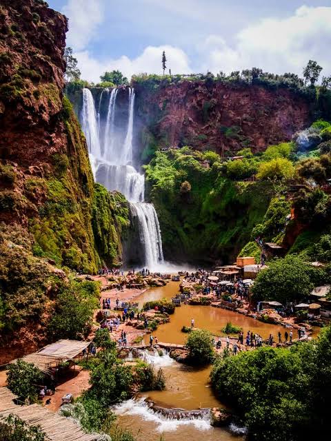 Spectacular Ouzoud Waterfalls in Morocco cascading down steep red rocky cliffs into a bustling river basin, surrounded by lush green vegetation, tourist boats, and scenic viewing paths.