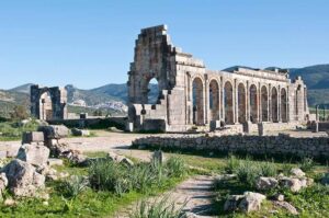 Ancient Roman ruins of Volubilis in Morocco featuring the historic stone arches of the Basilica, scattered archaeological ruins in the foreground, and scenic green mountains under a clear blue sky.