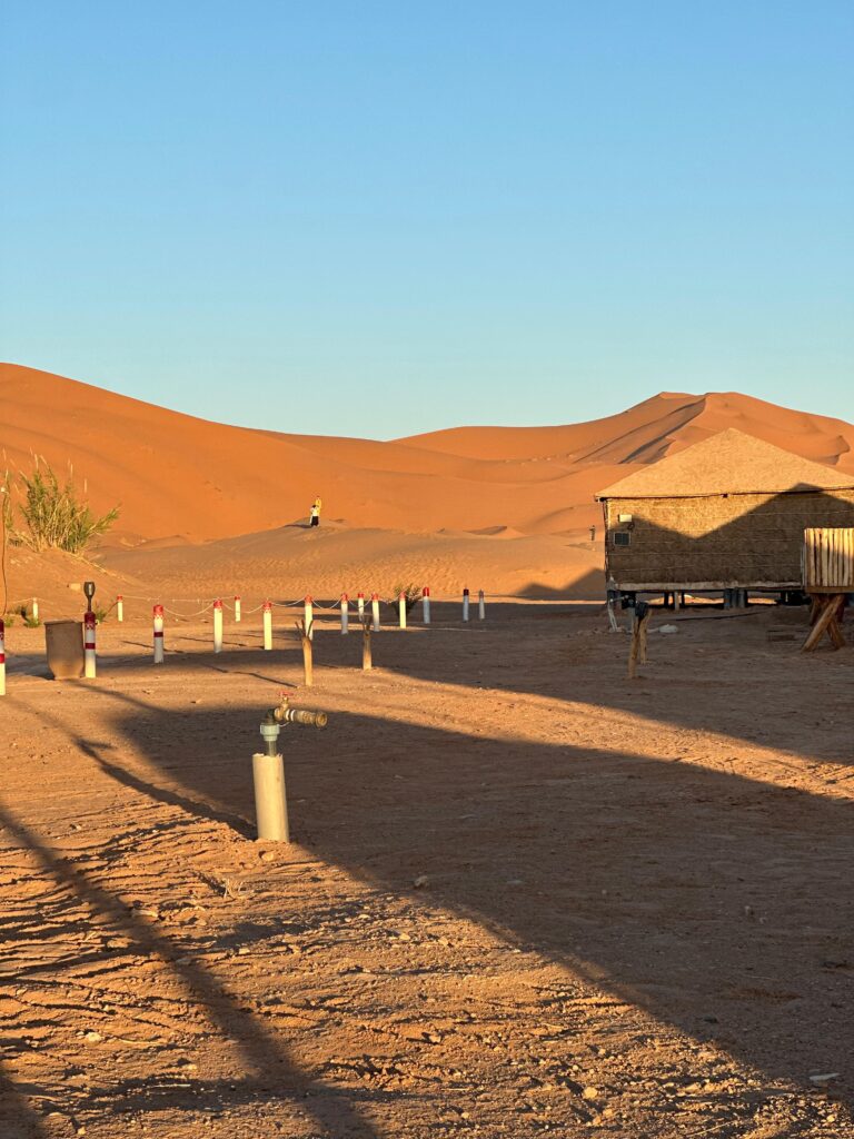 Desert camp landscape at sunset featuring glowing orange sand dunes, rustic architecture, long shadows, and a clear blue sky.
