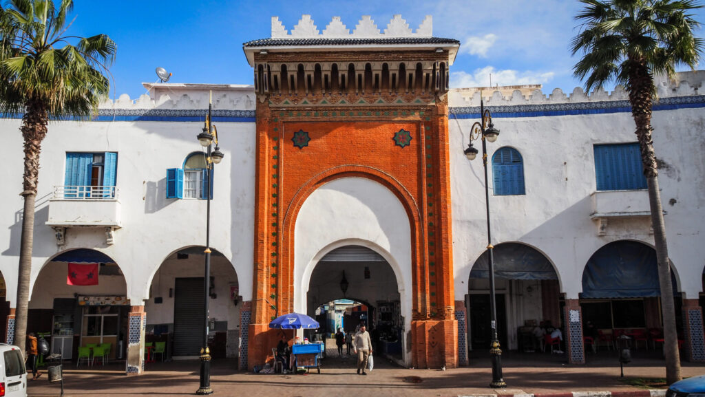 Historic entrance gate featuring a prominent orange-brick arch and white-washed arcade buildings lined with palm trees in Larache, Morocco.