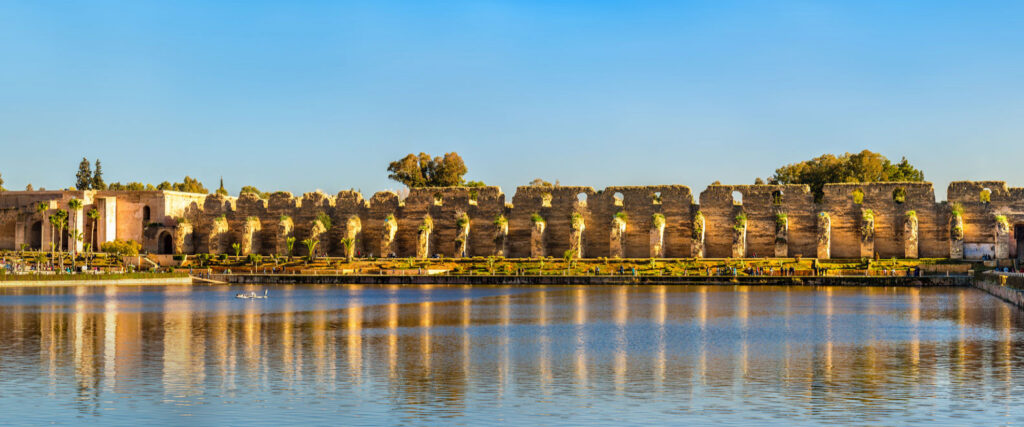 Scenic view of the historic Heri es-Souani granary and royal stables walls reflected in the tranquil waters of the Agdal Basin in Meknes, Morocco, under a clear sky.