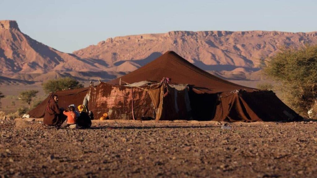 Traditional brown camel-hair Berber nomad tent pitched in a rugged Moroccan desert landscape, with a local family sitting together on the rocky ground against a mountainous backdrop.