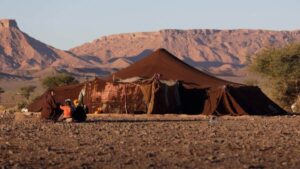 Traditional brown camel-hair Berber nomad tent pitched in a rugged Moroccan desert landscape, with a local family sitting together on the rocky ground against a mountainous backdrop.