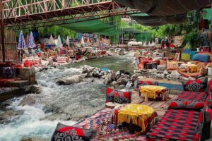 Scenic riverside dining area in the Ourika Valley, Morocco, featuring traditional low seating, colorful patterned cushions, and tables set up directly along the rushing mountain stream under shaded canopies.