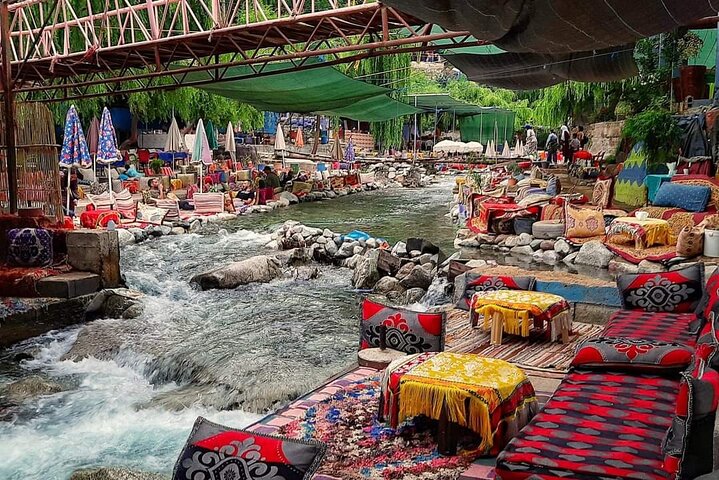 Scenic riverside dining area in the Ourika Valley, Morocco, featuring traditional low seating, colorful patterned cushions, and tables set up directly along the rushing mountain stream under shaded canopies.