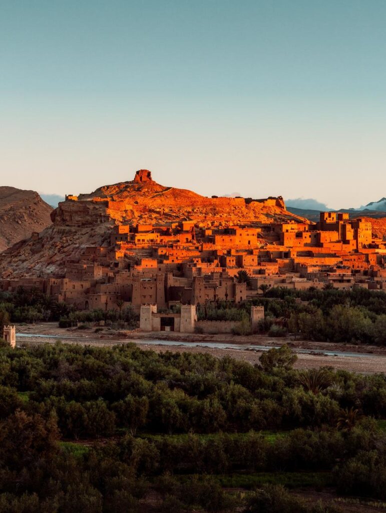 Historic fortified village of Aït Benhaddou in Morocco illuminated by glowing golden hour sunlight, featuring ancient earthen clay kasbah architecture built into a hillside, with a riverbed and lush green palm trees in the foreground.