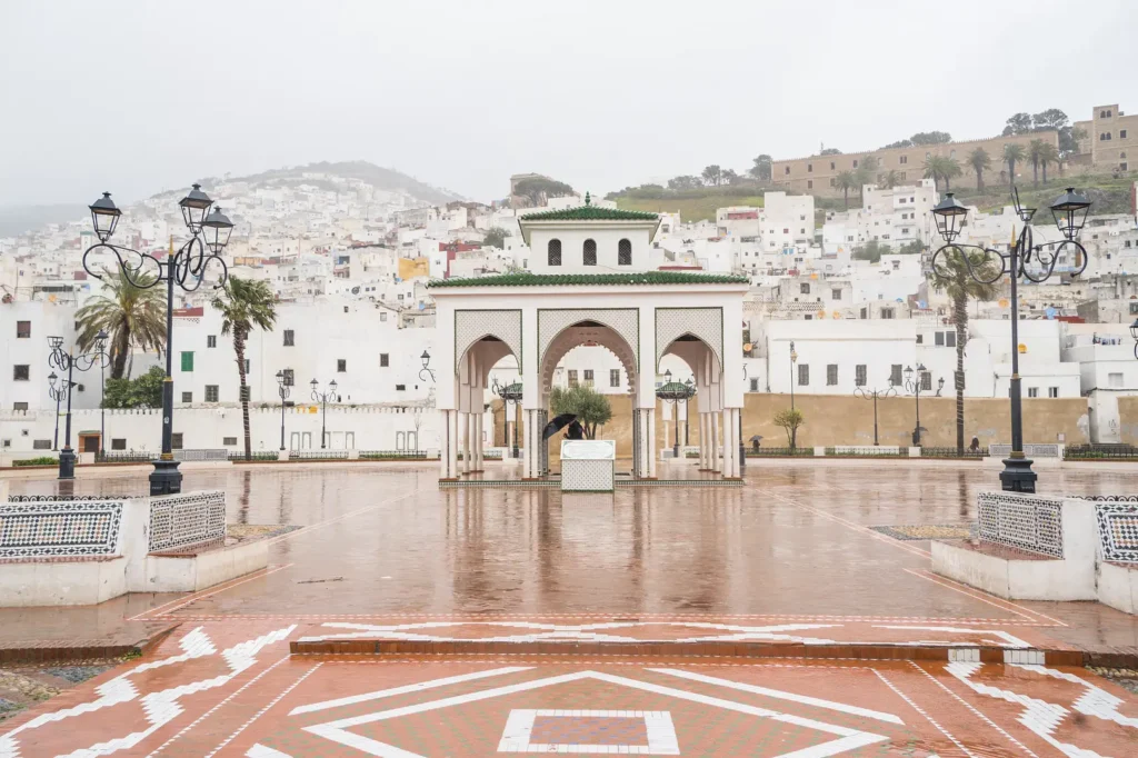 A rainy public square in Tetouan, Morocco, featuring an ornate white and green-tiled pavilion with traditional arches, surrounded by wet red-tiled paving and white-washed hillside buildings under an overcast sky.
