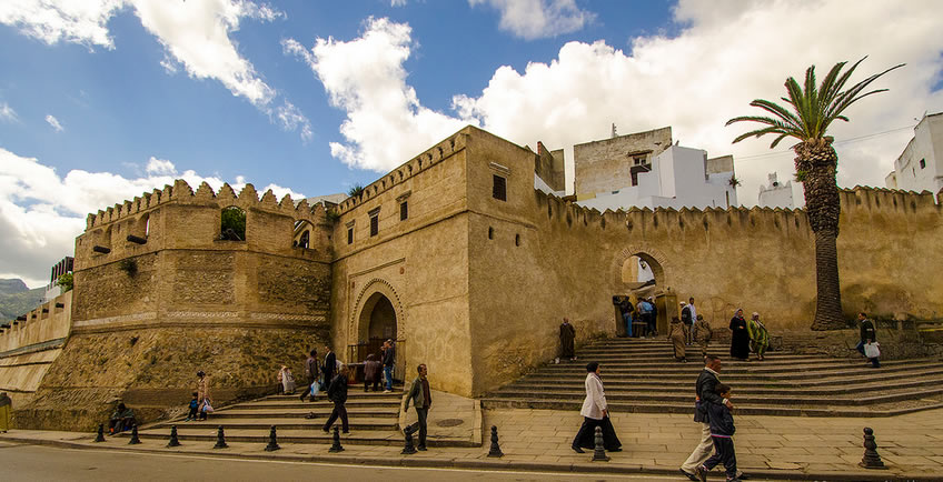 Historic stone ramparts and arched gateway of the old medina in Tetouan, Morocco, with people walking along the wide outdoor stone staircases and a tall palm tree under a dramatic blue and cloudy sky.