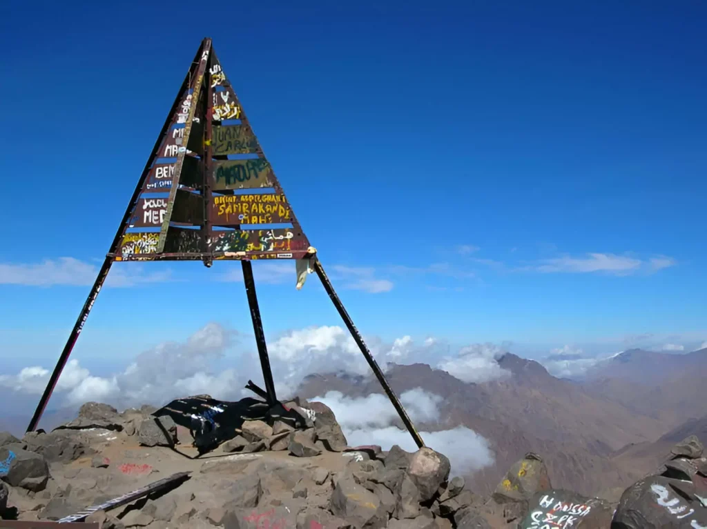 Iconic triangular metal summit marker on top of Mount Toubkal in Morocco, overlooking a vast panoramic view of sea of clouds and the surrounding High Atlas mountain range under a clear blue sky.