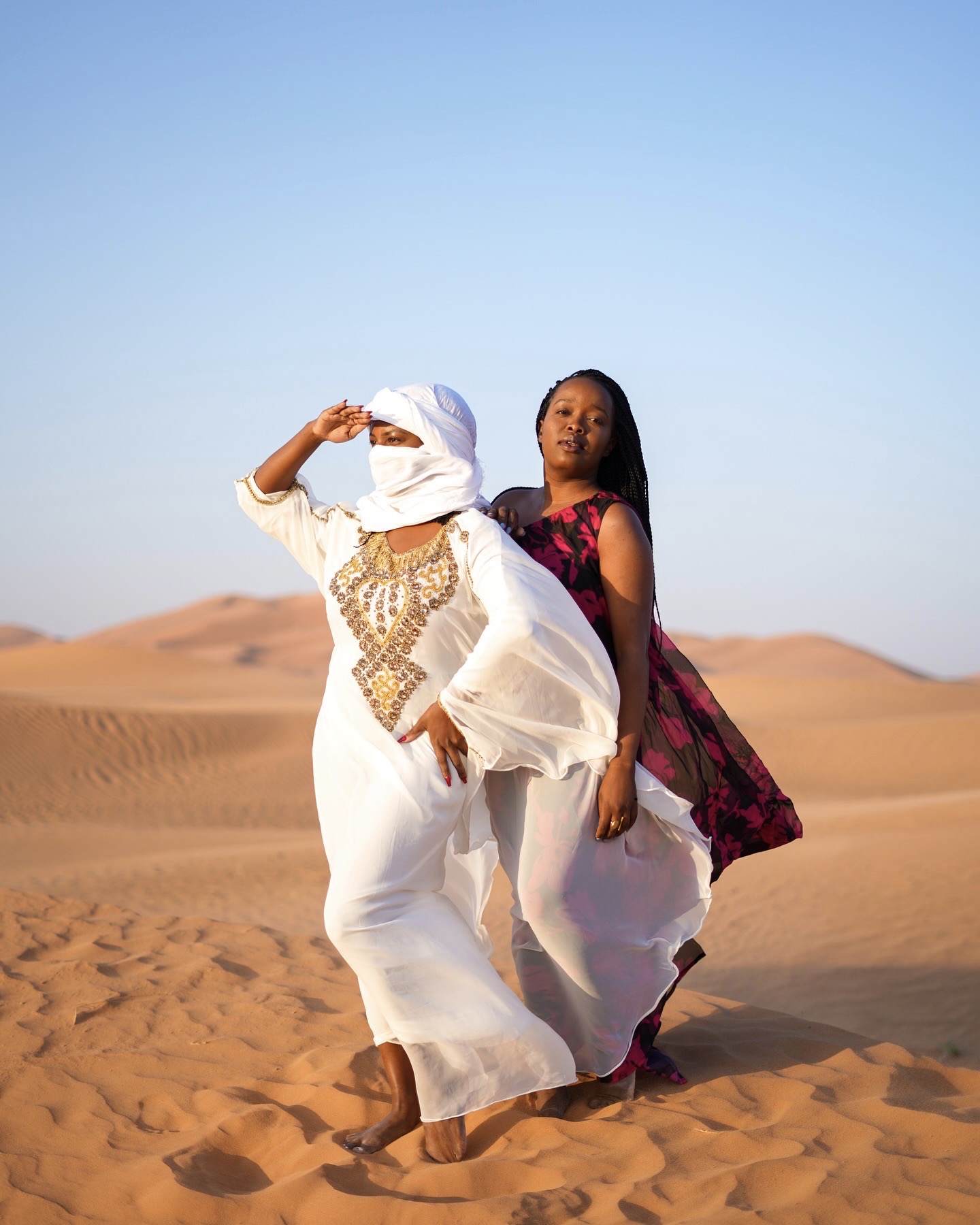Two women in long white dresses and scarves on the Sahara dunes