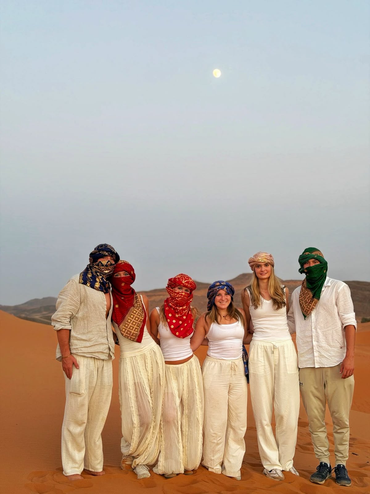 Group in white cotton trousers and colorful scarves on a Sahara dune at dusk