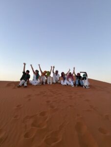 Nine students sitting on a Sahara dune with their arms raised in colorful desert scarves