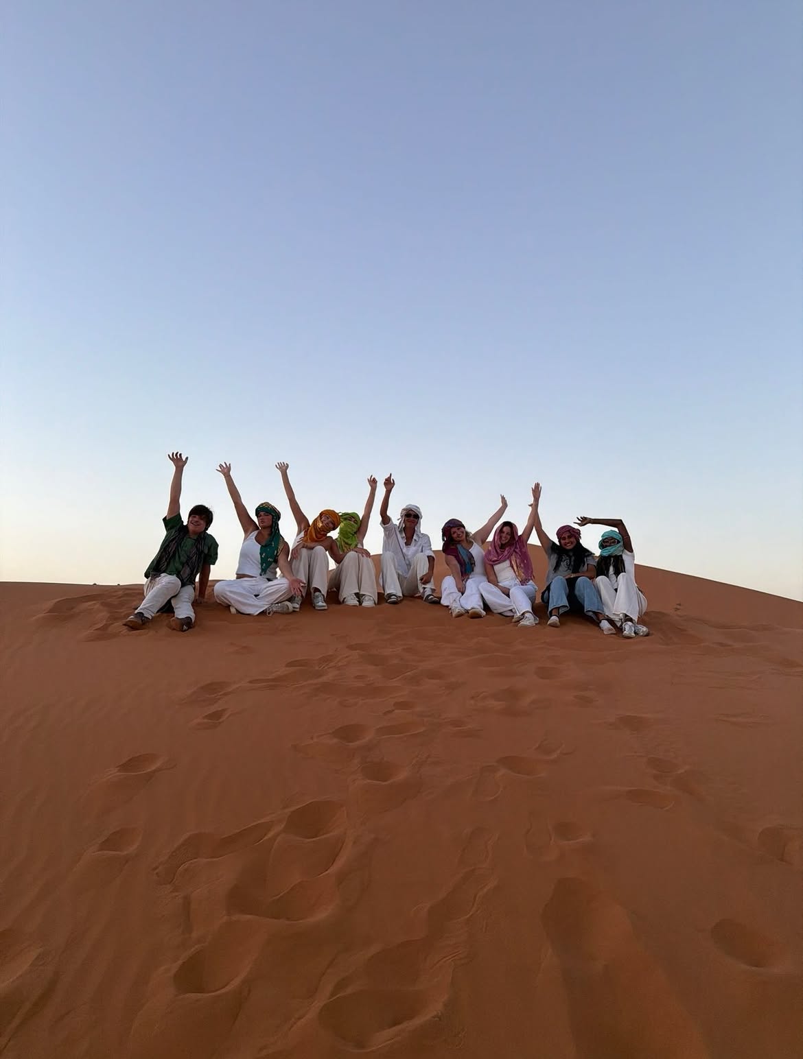 Nine students sitting on a Sahara dune with their arms raised in colorful desert scarves