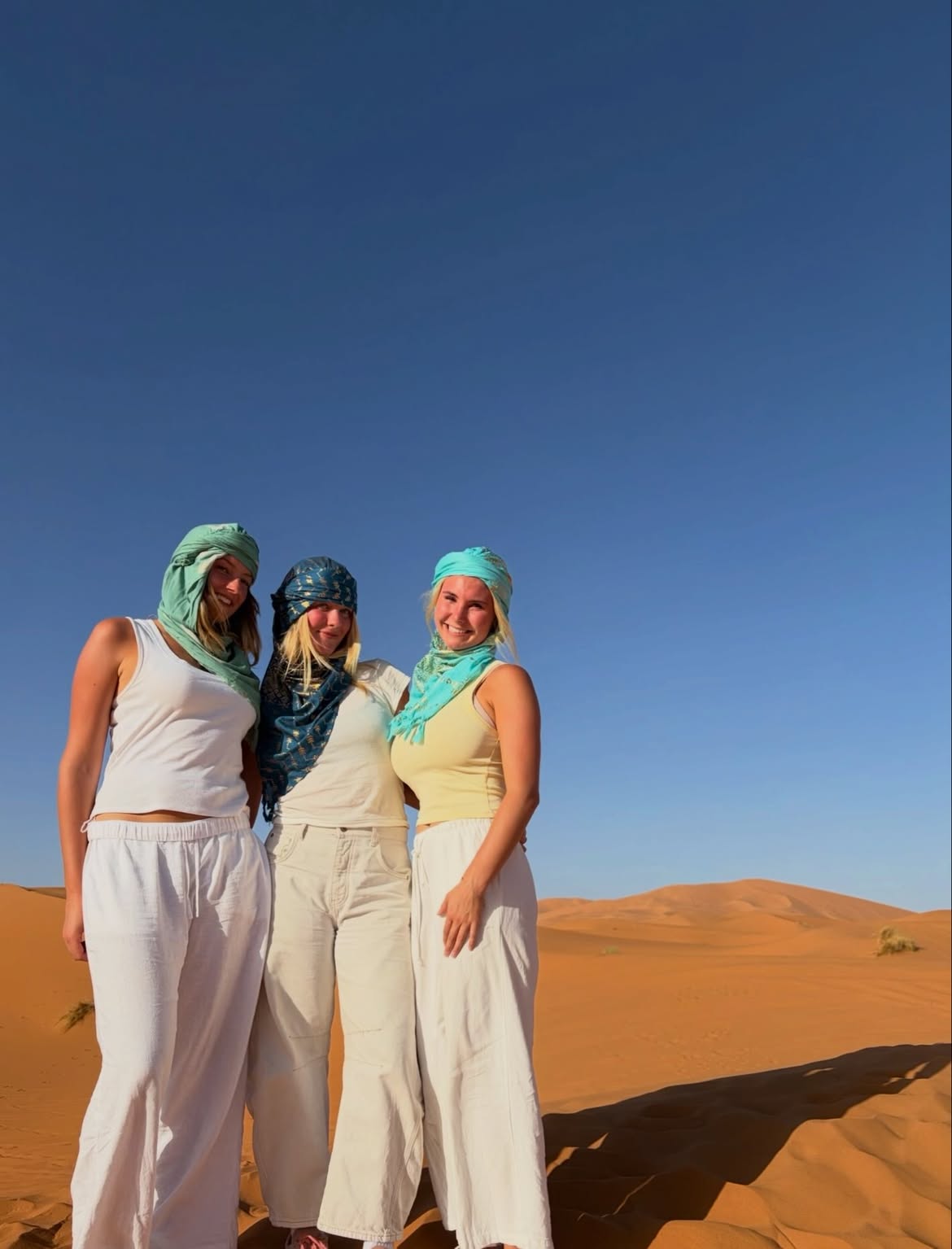 Three friends in desert scarves smiling on a Sahara dune under a deep blue sky