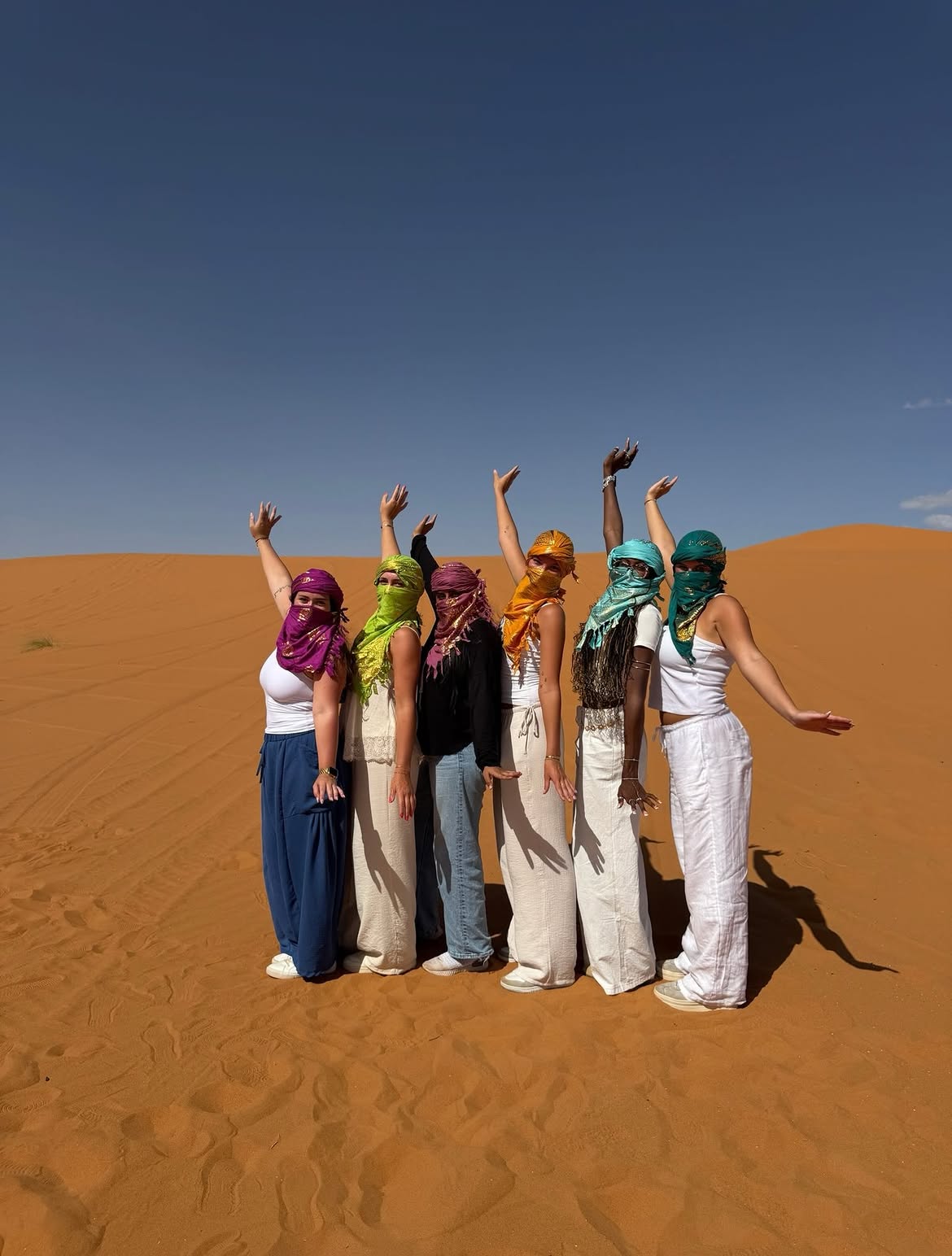 Six students in colorful face scarves with raised arms on a Sahara dune