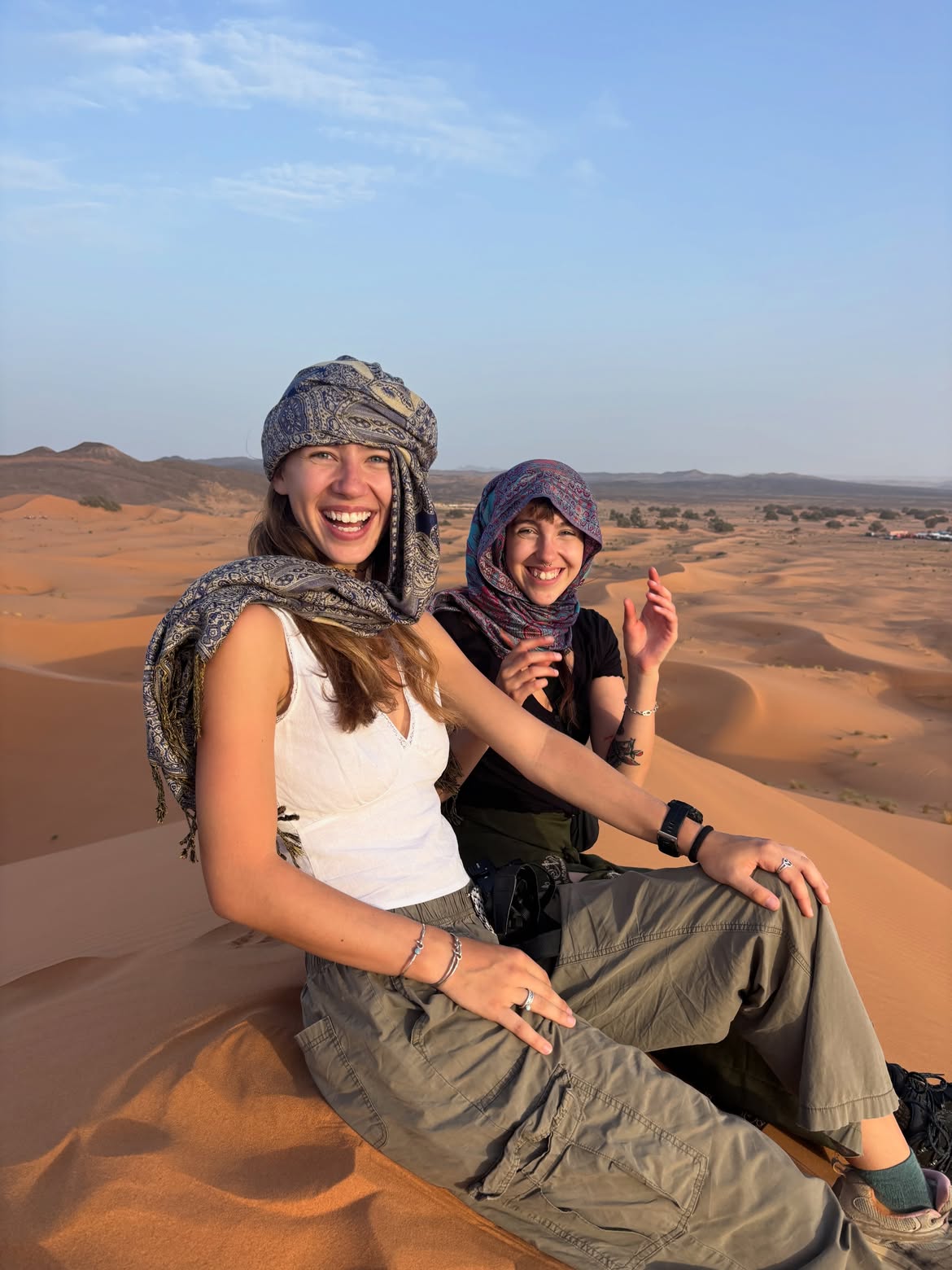 Two friends laughing in desert scarves on a Sahara dune at sunset