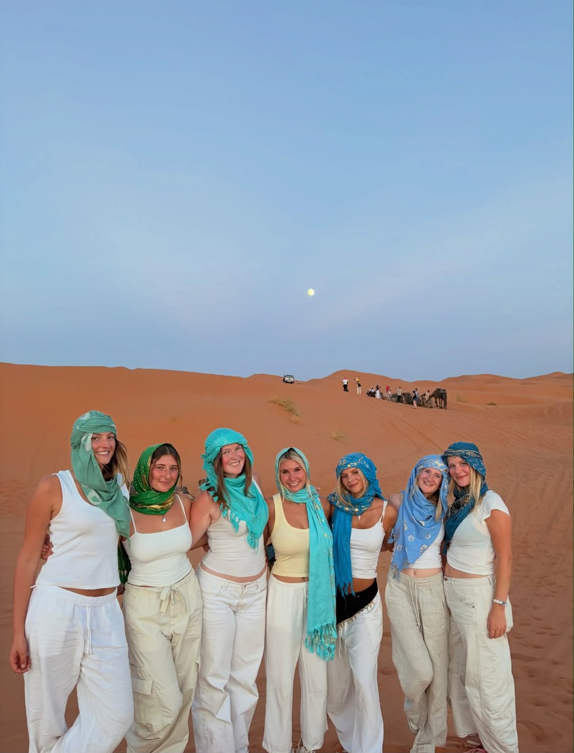 Six students in desert scarves standing on a Sahara dune at dusk with a camel caravan behind