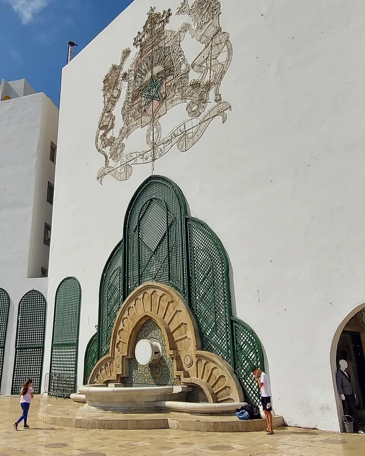 Green-tiled fountain and the Moroccan coat of arms on a white wall in a Tetouan square