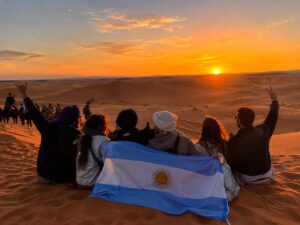 Grupo de viajeros argentinos con la bandera de Argentina mirando el atardecer en las dunas del Sahara