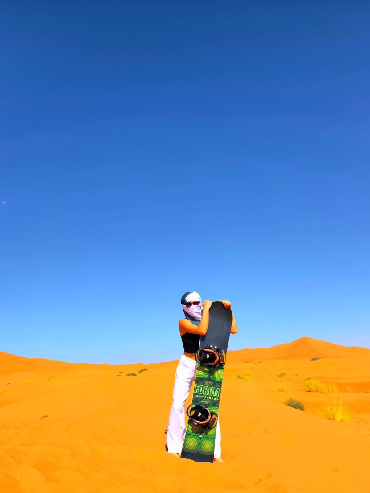 Traveler holding a sandboard on a Merzouga dune under a clear blue sky